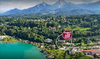 A picturesque view of Lake Faak with surrounding mountains and a green landscape. In the foreground, the Faaker See Inn Hotel is clearly visible.