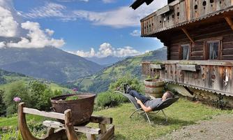A cozy man relaxes on a lounge chair in front of an Alpine cabin. In the background, a beautiful mountain landscape stretches out under a blue sky.