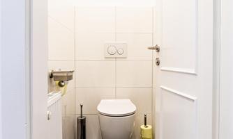 A simple, modern restroom with white tiles and a black floor. The doors are light, and there is a toilet paper holder as well as a toilet brush holder.