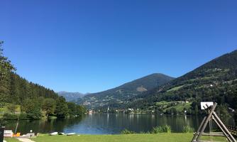 A tranquil lake surrounded by mountains and forests. The sky is clear and blue, perfect for a relaxing day in nature.