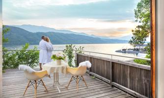 A couple in bathrobes stands on a terrace overlooking the lake. The surroundings are calm and picturesque, surrounded by mountains and trees.