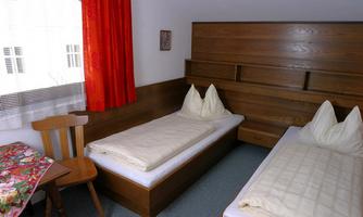 A simple guest room with two single beds and a table. The walls are in warm wood tones and the window has a white curtain.