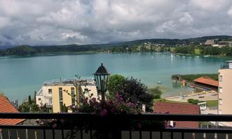 A picturesque view of a clear lake with green shores and gentle waves. In the foreground, houses and blooming plants can be seen.