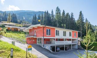 A modern building in an idyllic mountain landscape, surrounded by trees and meadows. The sun shines clearly in the sky and there is an asphalted path leading to the building.