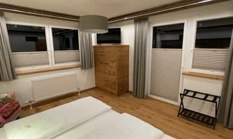 A bright bedroom with two single beds. Large windows with curtains and a wooden wardrobe are also visible.