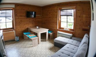 A cozy living space with wooden walls and large windows. The room contains a sofa, a table, and a television.