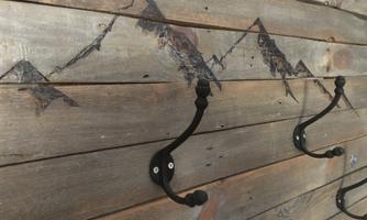 A rustic wooden wall with a mountain painting. Two black hooks are hanging on the wall.