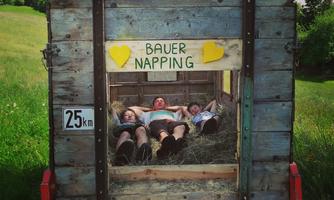 Three people are lying relaxed in a wooden trailer on hay. Above them hangs a sign with the inscription "FARMER NAPPING".