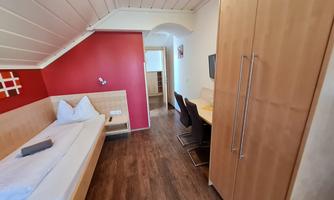 A modern room with a wooden floor and a red accent wall. There is a bed, a table with chairs, and a television.