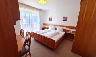 A bright room with a double bed and wooden furniture. There is a table with chairs and large windows that let in plenty of light.