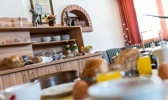 A covered breakfast table with various foods and drinks. In the background, there is a wooden shelf with more groceries visible.