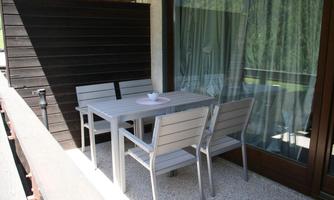 A small balcony with a table and four chairs. The furniture is made of light material and is next to a large front window.