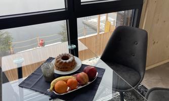 A beautiful table with a fruit bowl and a bundt cake in a bright room. In the background, there are large windows that offer a view outside.