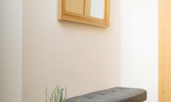 A bright entrance area with a gray bench and a round flower pot. A wooden frame with a mirror hangs on the wall.