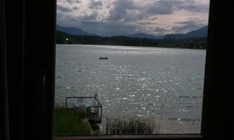 A tranquil lake with shimmering water and a cloudy sky. In the foreground, a window is visible that opens up the view of the landscape.