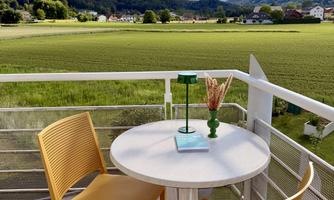 A cozy balcony with two yellow chairs and a white table. In the background, green meadows and mountains can be seen under a blue sky.