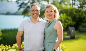 A couple is smiling outdoors in front of a lake. Trees and a green landscape can be seen in the background.