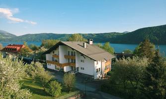 A beautiful house near a lake, surrounded by green trees and blooming plants. In the background, a picturesque landscape with mountains can be seen.