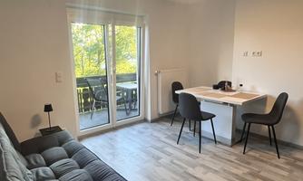 A modern living room with a sofa bed and a table with chairs. Large windows provide plenty of daylight and a view of the balcony.