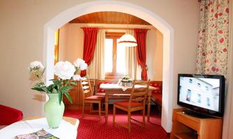 A cozy room with a dining area and a television. There are flowers in a vase on the table and the walls are fitted with curtains.