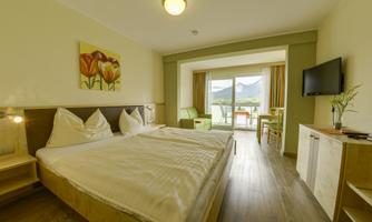 A modern bedroom with a large double bed and bright furniture. The room has access to a balcony with a view of the mountains.