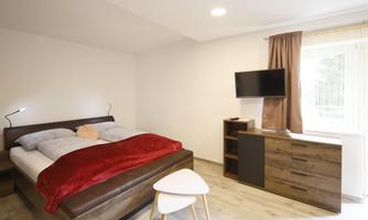 A modern bedroom with a large bed, equipped with red and blue bedding. Next to the bed is a small table and a TV on the wall.