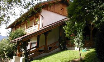 A charming house with a wooden base and a balcony. Surrounded by green grass and trees, it radiates coziness.