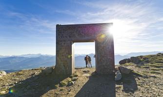 A large gate stands on a mountain peak, surrounded by an impressive mountain landscape. Two people stand in front of it, enjoying the view.