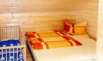 A cozy bedroom with wooden paneling. There is a double bed with colorful pillows and a crib next to it.