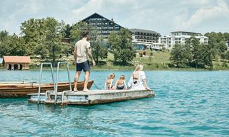 A family is sitting on a pier and looking at the water. In the background, trees and a hotel are visible.