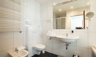 A modern bathroom with white tiles and a clear mirror wall. There is a sink, a toilet, and a towel warmer.