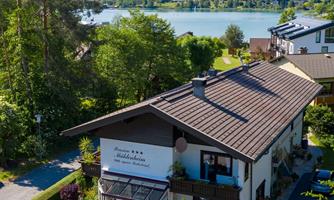 A picturesque house by the water with a view of the mountains. Surrounded by lush greenery and a clear sky.