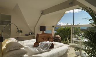 A modern bedroom with a large window and a view of the outdoors. There is a bed, a table, and some plants.