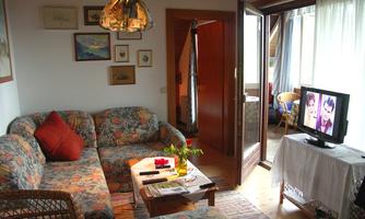 A cozy living room with a colorful sofa and a low table. The TV shows pictures and there are large windows with plenty of light.