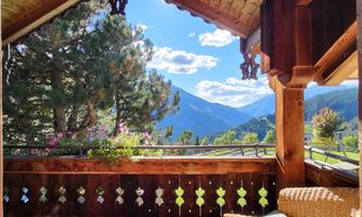 A beautiful balcony with wooden cladding and a view of the mountains. In the background, green trees and a blue sky with a few clouds can be seen.