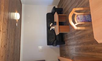 A simple living room with a black sofa and a wooden table. The room has a wooden floor and some wooden furniture pieces.