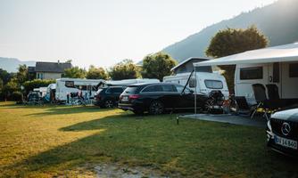 A campsite with several motorhomes and cars. In the background, green hills and a clear sky can be seen.