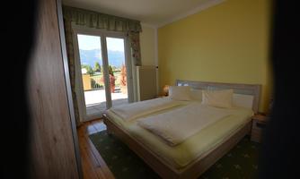 A cozy bedroom with a large bed and decorative pillows. Windows open to a terrace with a view of the mountains.