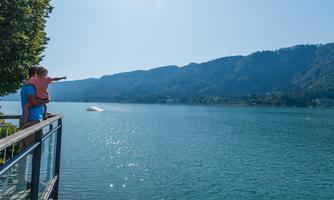 A father is holding his child on his shoulders and pointing at a lake. In the background, green hills and a clear blue sky can be seen.