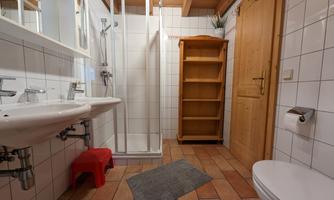 A modern bathroom with a shower, two sinks, and a wooden shelf. The walls are tiled, and there is a bright wooden beam.