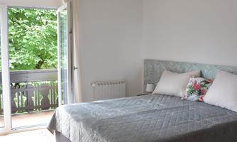 A bright bedroom with a large bed and a cozy atmosphere. The view looks out onto a balcony with green trees in the background.