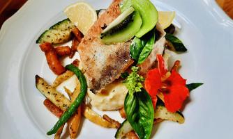 An attractively arranged plate with fried fish, colorful vegetables, and fresh herbs. The dish is decorated with lemon slices and edible flowers.