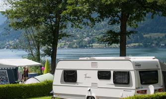 A caravan is parked at a campsite by the lake, surrounded by trees. In the background, green mountains and a picturesque village can be seen.