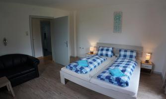 A bright bedroom with a double bed and checked bedding. There is a cozy armchair and subtle wall decoration.