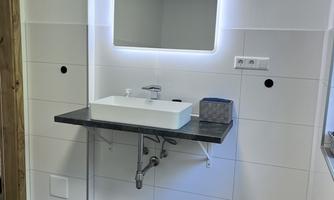 A modern bathroom with a floating sink and a lighted mirror. The floor is tiled with gray tiles and a black bath mat is placed in front of the sink.