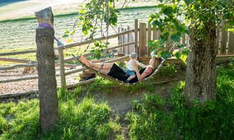A woman is relaxing in a hammock and holding a child in her arms. Surrounded by trees and a green meadow, the scene exudes tranquility and harmony.