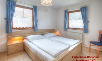A bright bedroom with two single beds and blue curtains. The room has two windows and a cozy atmosphere.