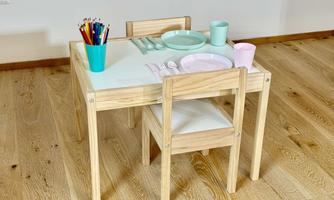 A children's table made of wood with two chairs. On the table are colorful pens and dishware in pastel colors.