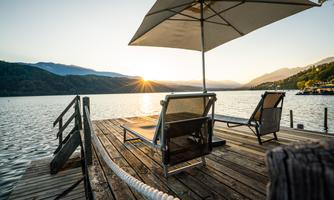 A quiet terrace by the water with two sun loungers and a beach umbrella. The sun sets behind the mountains and reflects on the water's surface.