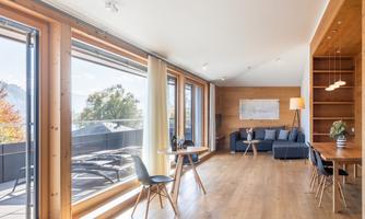 A modern living room with large windows that let in plenty of light. The wooden floor and furniture give the room a cozy atmosphere.